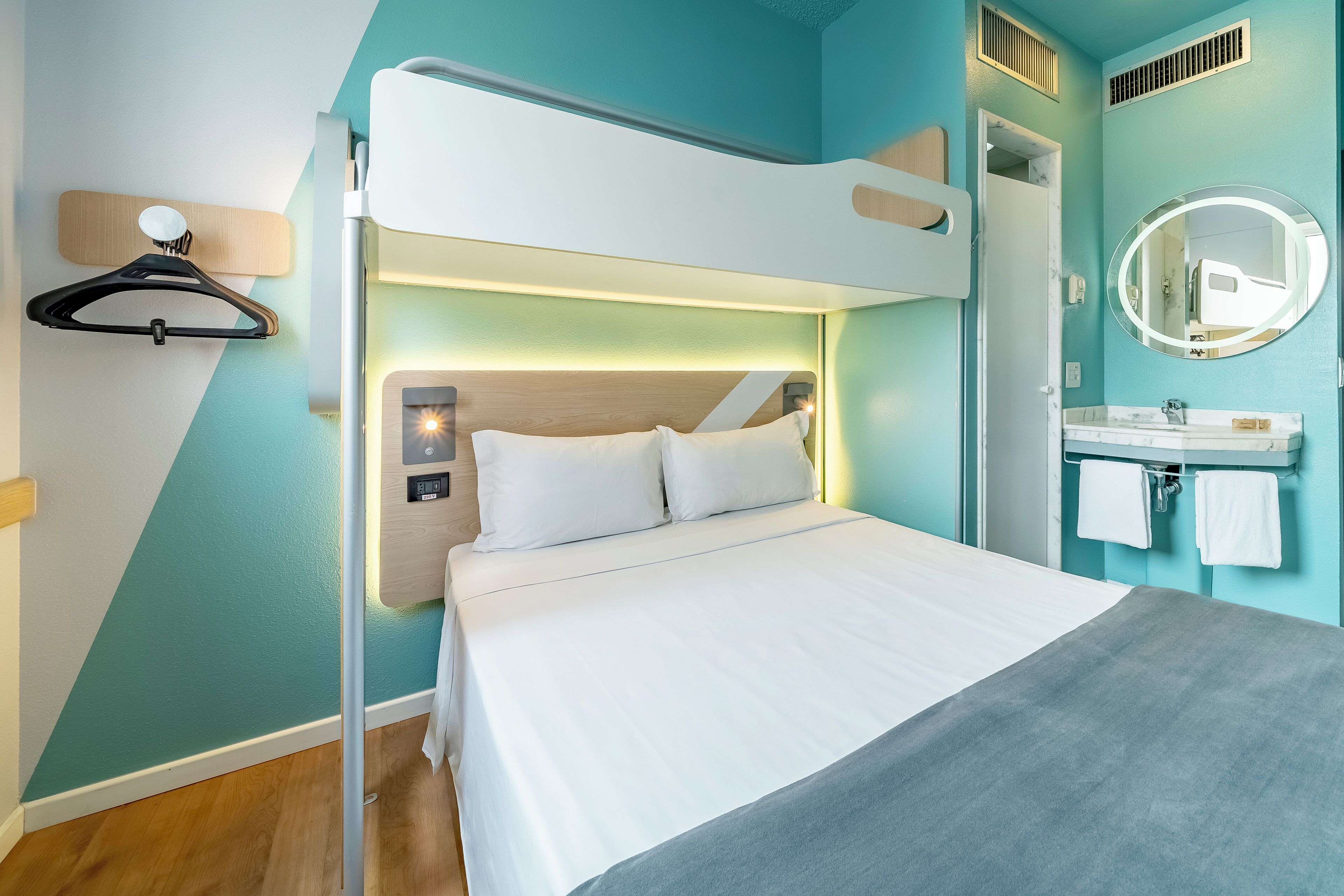 Een moderne hotelkamer met een stapelbed, een nachtkastje met een lamp, een ronde spiegel met verlichting, en een badkamer met een wastafel en handdoekhouder.
