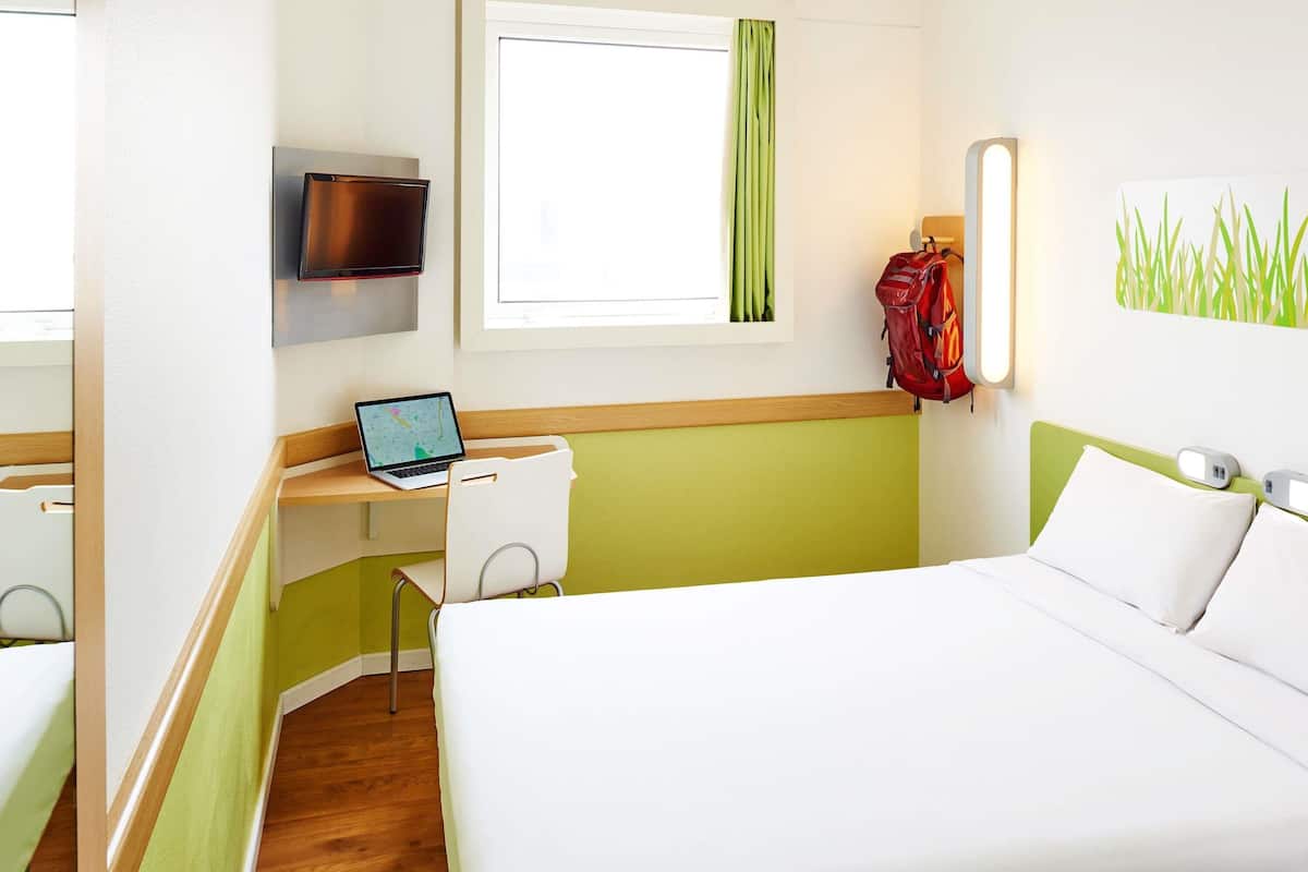A hotel room with a bed, a desk with a laptop, a TV, and a red backpack hanging on the wall.