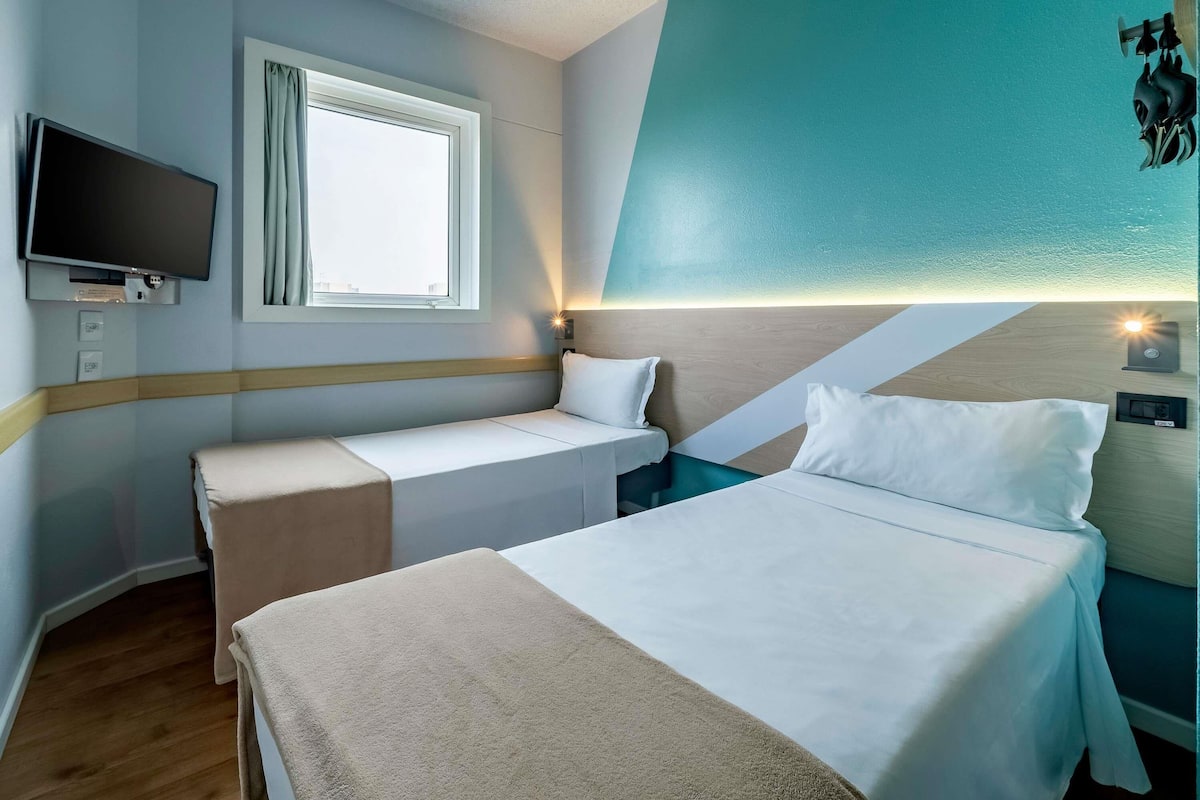 A hotel room with two beds, a TV, a window with curtains, and a wall-mounted light.