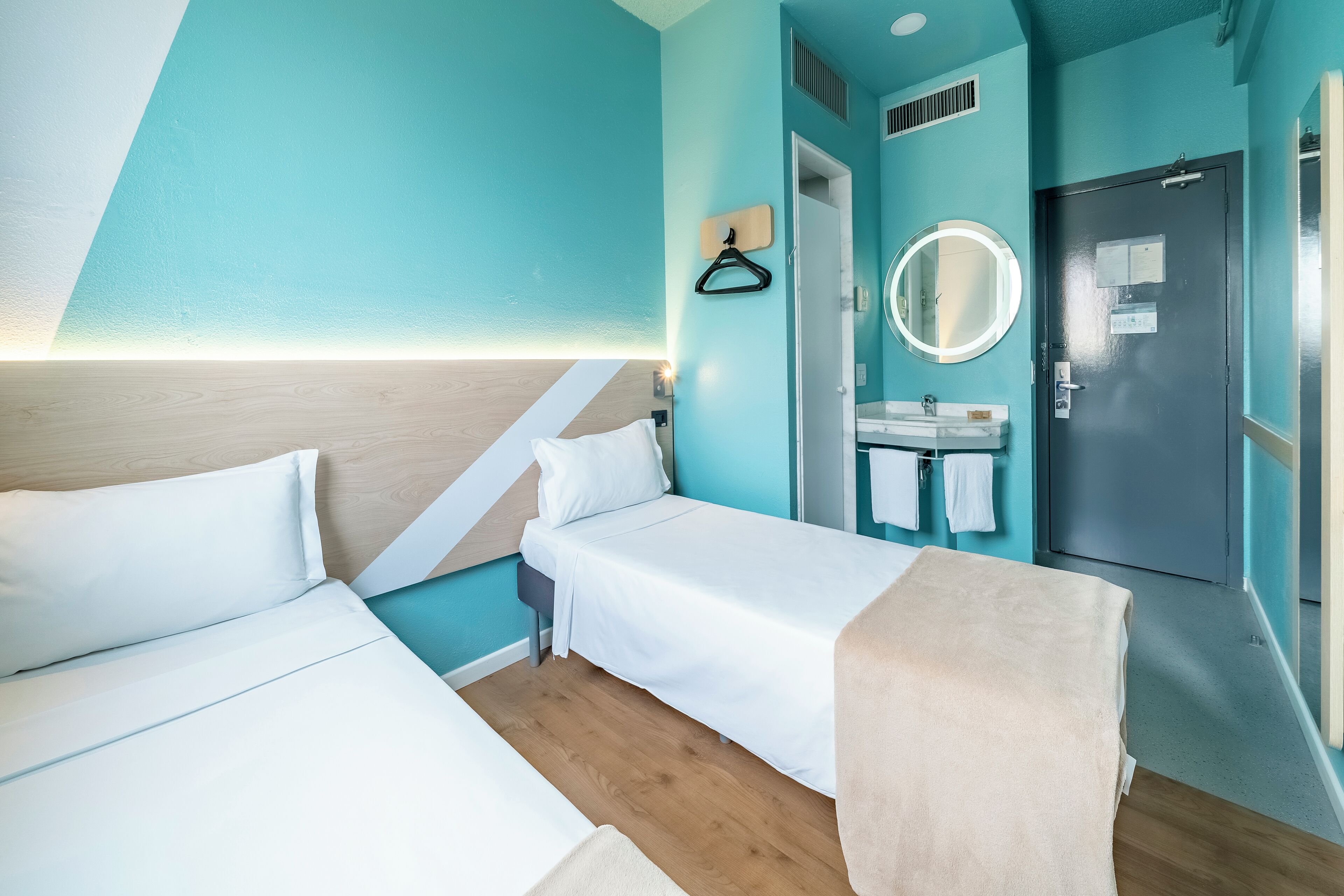 Habitación de hotel con dos camas, un baño con lavabo y espejo, y una puerta con perchero para toallas.
