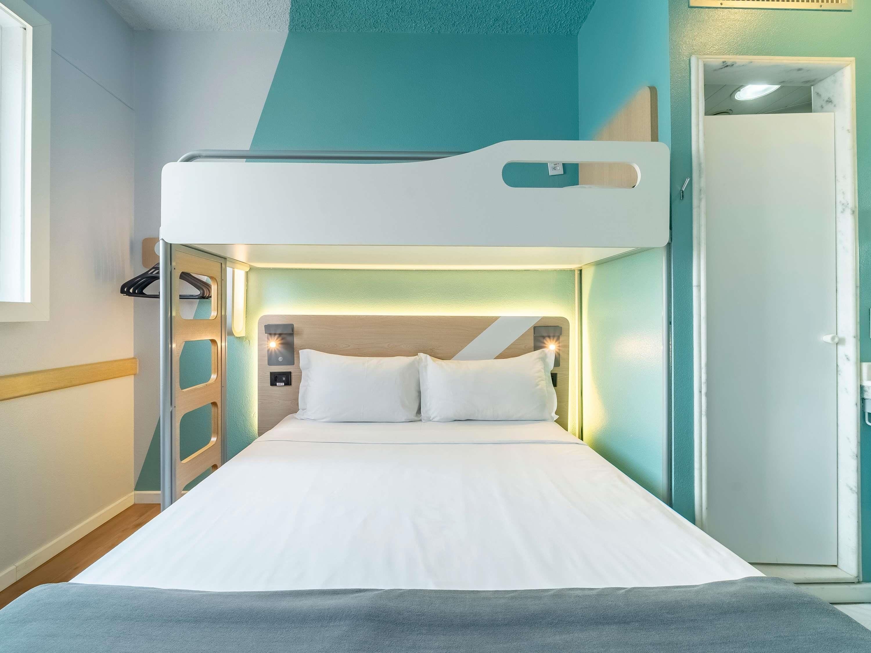 Uma cama beliche com colchão branco, cabeceira com iluminação integrada e uma porta à direita.