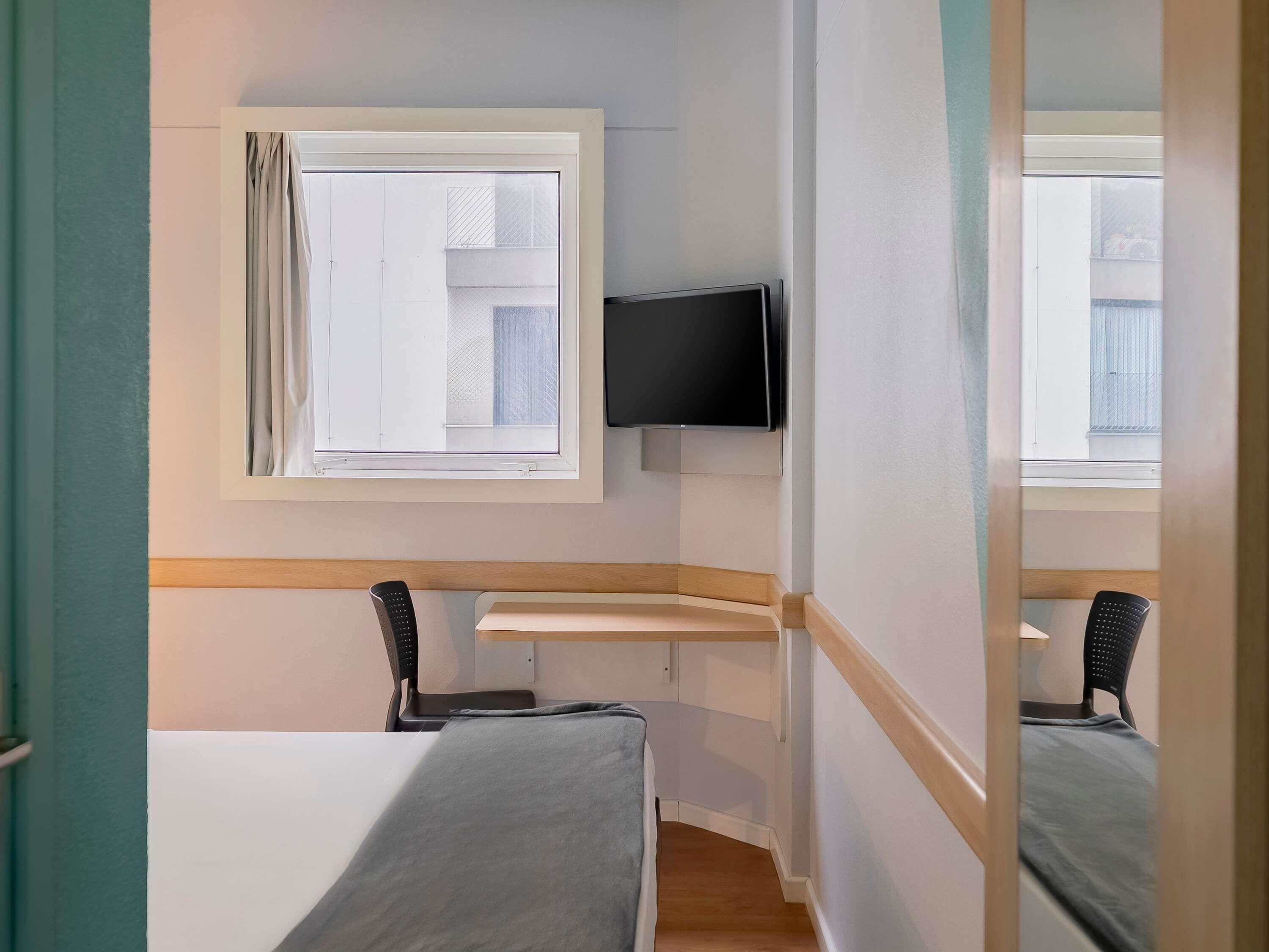A hotel room with a bed, a desk, a chair, a television, and a window.