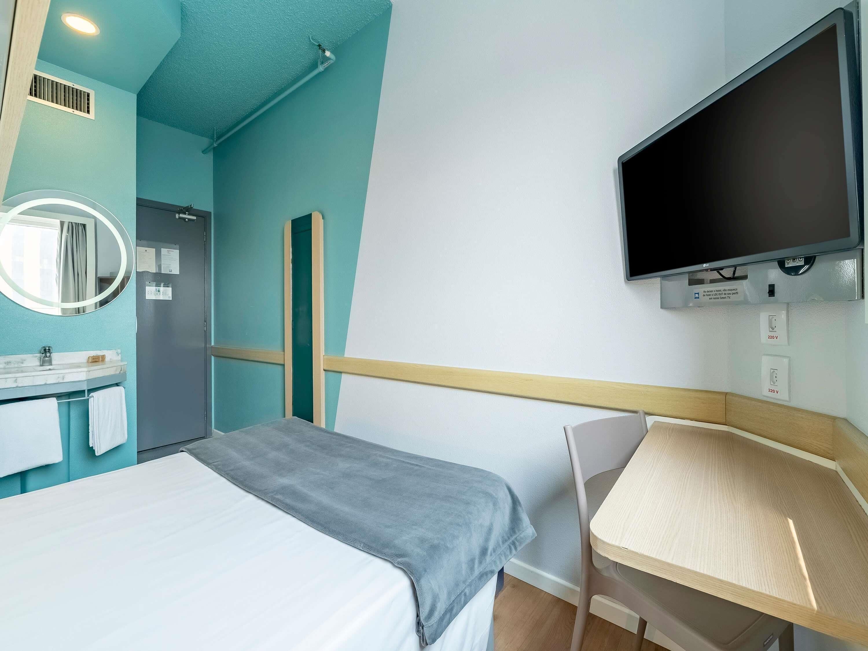 Quarto de hotel com cama, escrivaninha, televisão, espelho e banheiro com pia e porta-toalhas.
