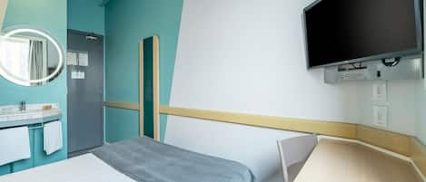 Quarto de hotel com cama, escrivaninha, televisão, espelho e banheiro com pia e porta-toalhas.