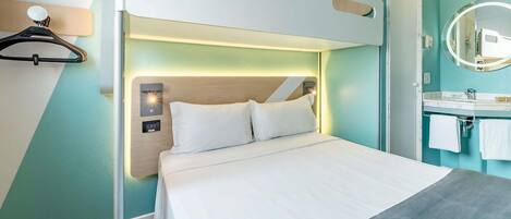 Una habitación de hotel moderna con una cama litera, una mesita de noche con lámpara, un baño con lavamanos y espejo, y paredes color turquesa.