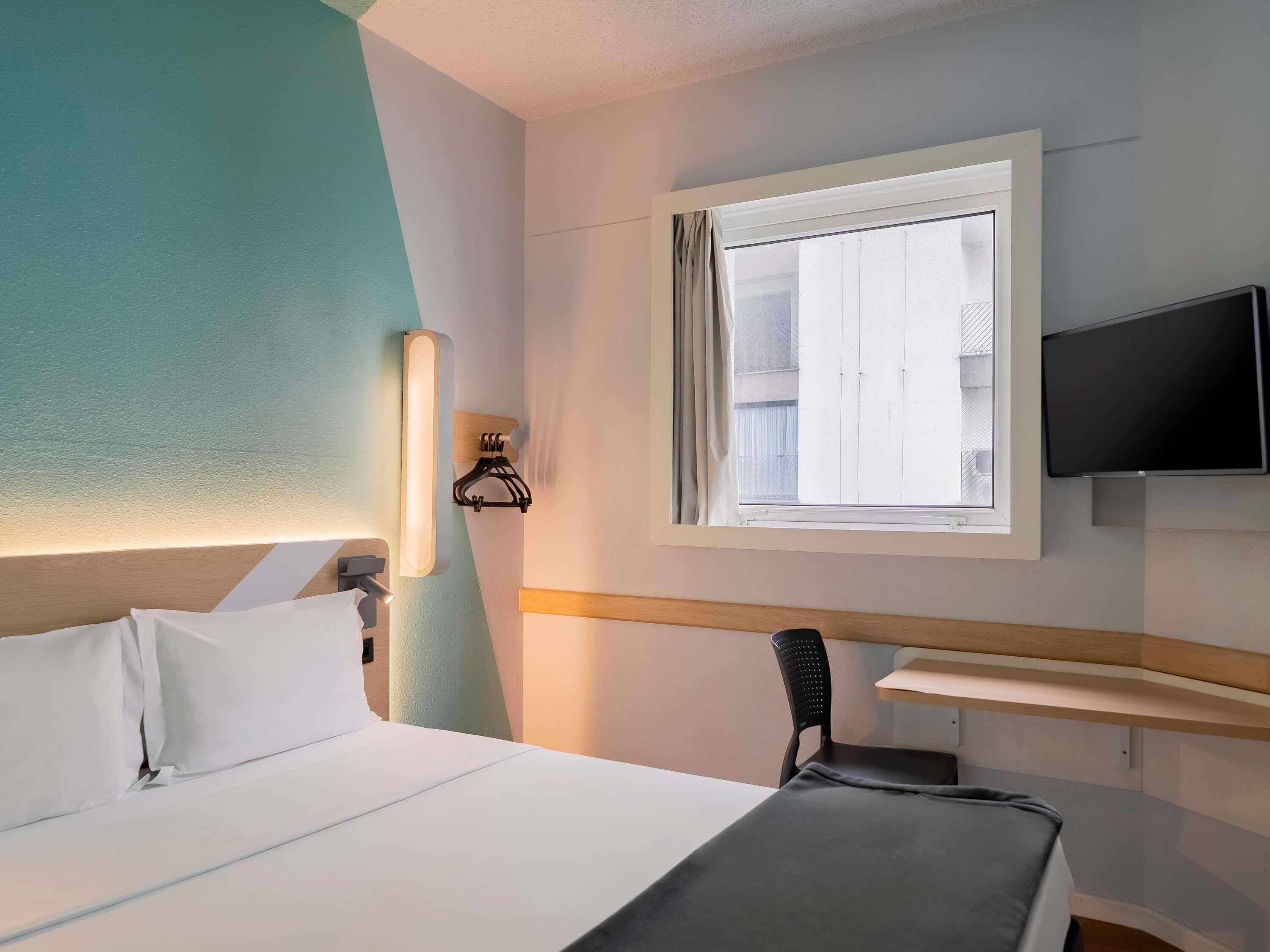 A hotel room with a bed, a TV mounted on the wall, a window with curtains, and a desk with a chair.
