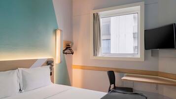 A hotel room with a bed, a TV mounted on the wall, a window with curtains, and a desk with a chair.