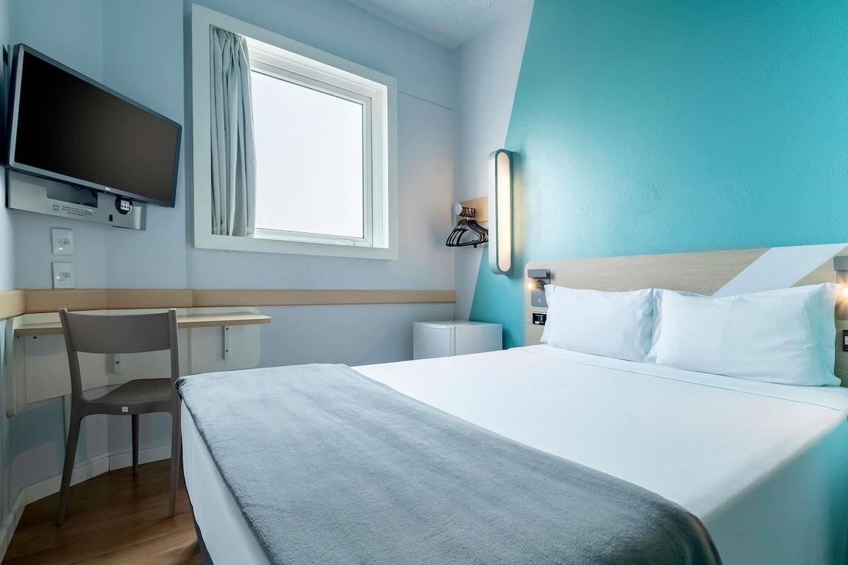 A hotel room with a bed, a desk, a chair, a television, and a window.