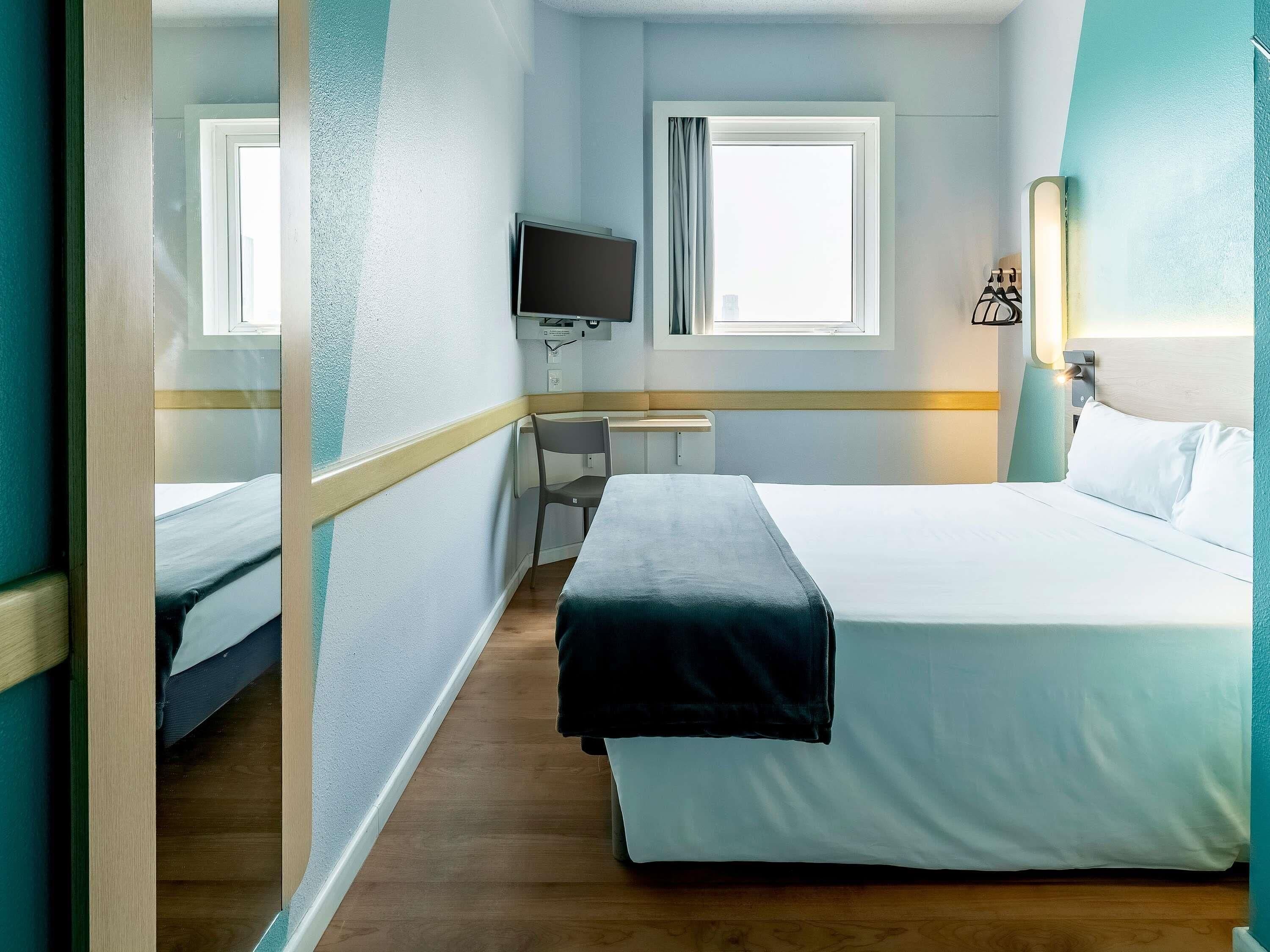 Quarto de hotel com cama, escrivaninha, cadeira, televisão e janela.