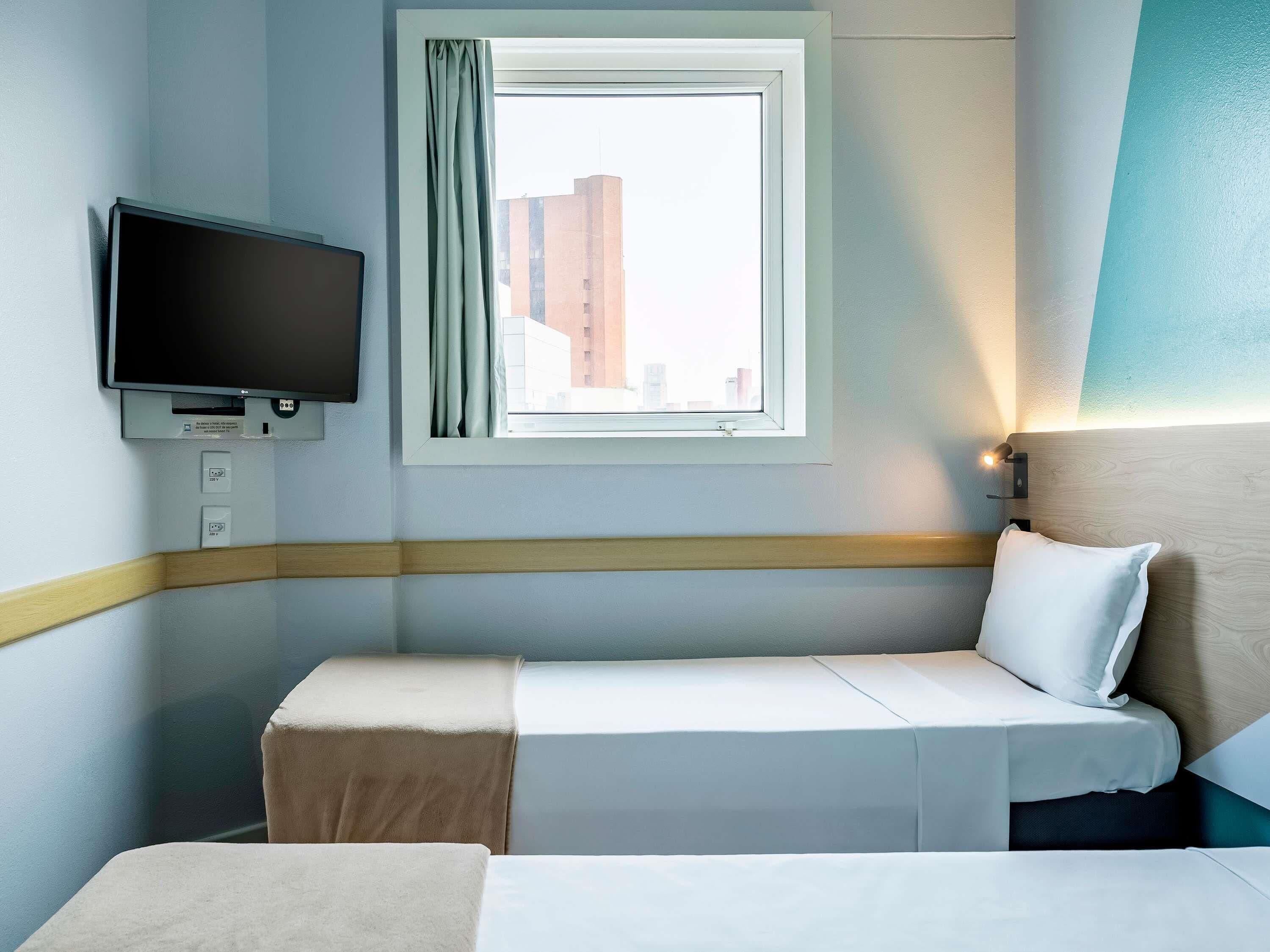 Habitación de hotel con cama, televisor montado en la pared, ventana con cortinas y vista a edificios.