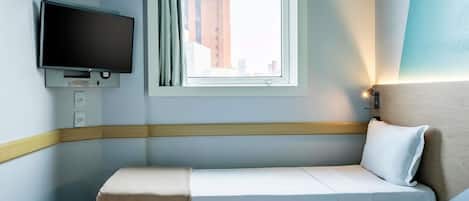 Habitación de hotel con cama, televisor montado en la pared, ventana con cortinas y vista a edificios.