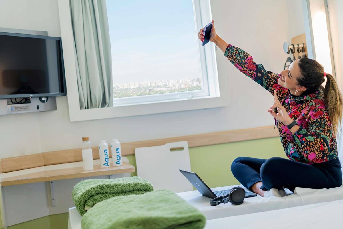 Una habitación con cama, una computadora portátil, auriculares y una ventana con vista a edificios.
