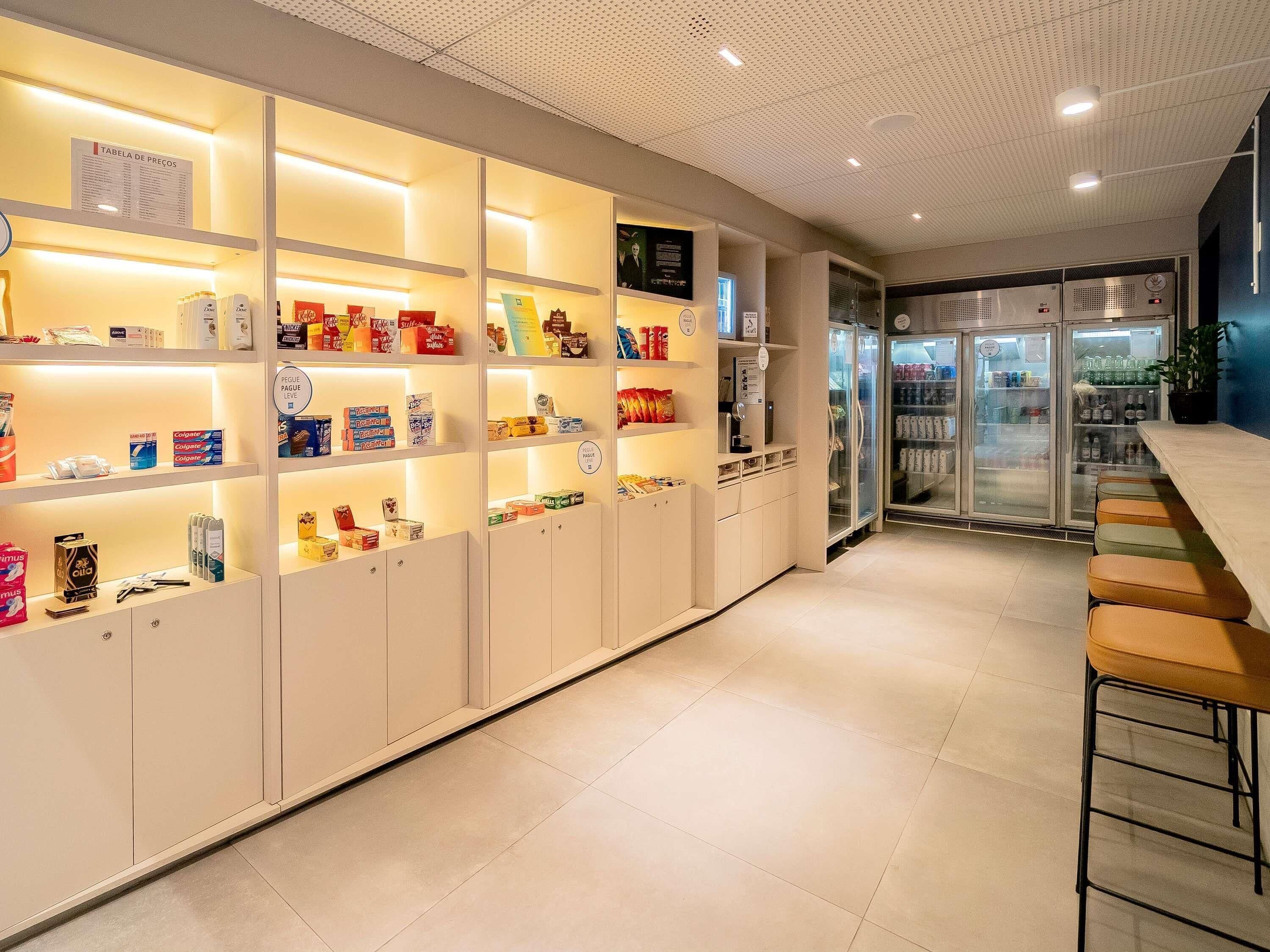 Un moderno vestíbulo de hotel con un mostrador de recepción alargado, un expositor refrigerado y diversos productos en estanterías.