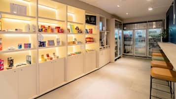 Un moderno vestíbulo de hotel con un mostrador de recepción alargado, un expositor refrigerado y diversos productos en estanterías.