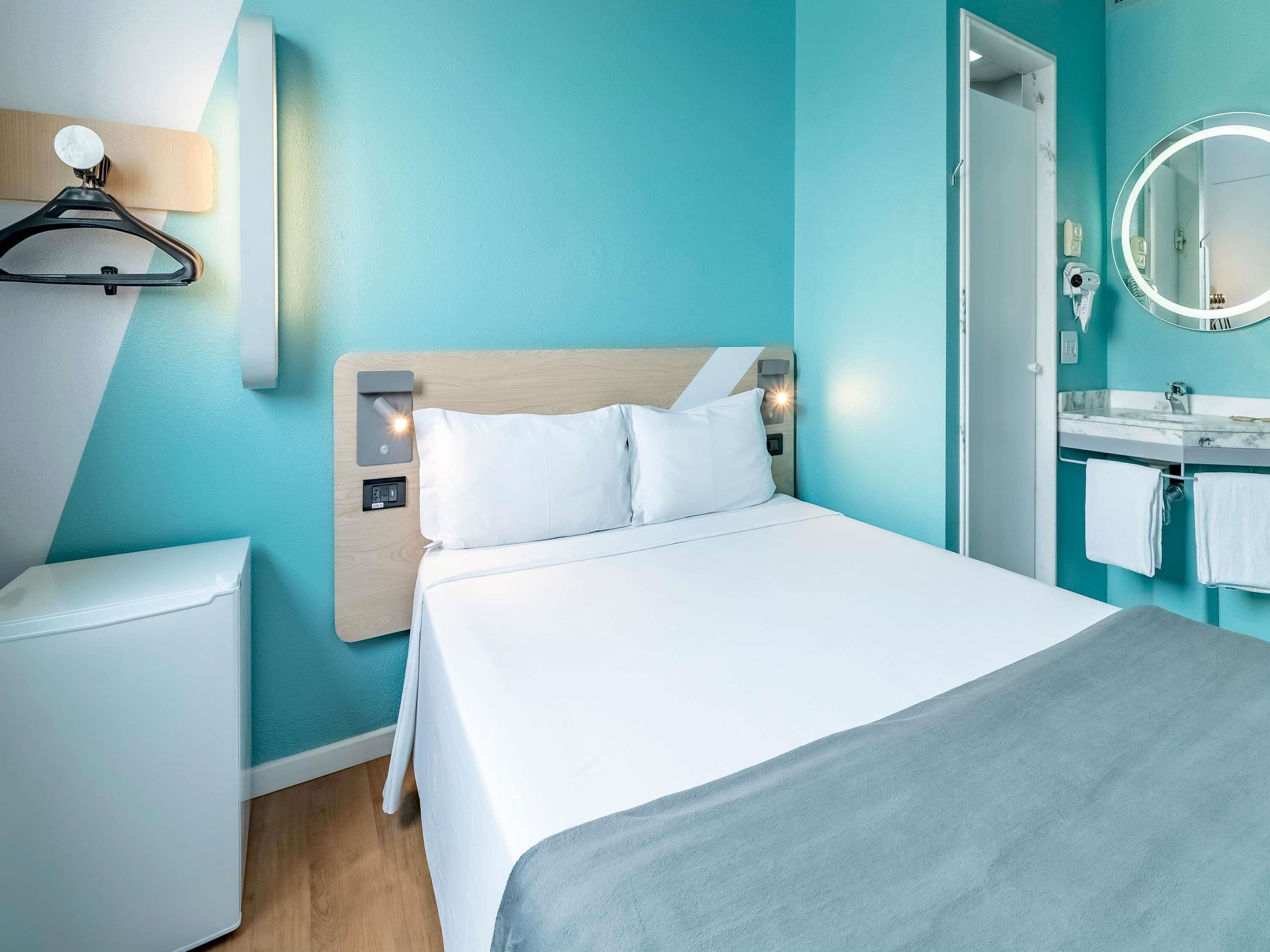 Een moderne hotelkamer met een bed, nachtkastje, wandlamp, spiegel en een badkamer zichtbaar door een geopende deur.