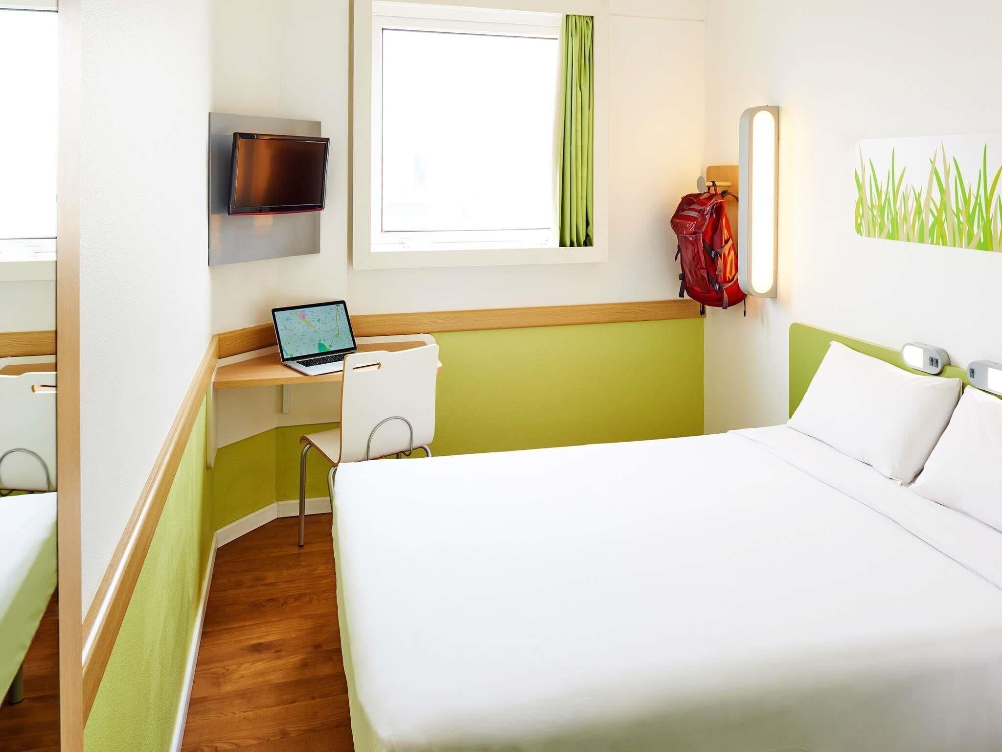 Et hotelværelse med en seng, et skrivebord med en bærbar computer, et fjernsyn og en rød rygsæk hængende på væggen.