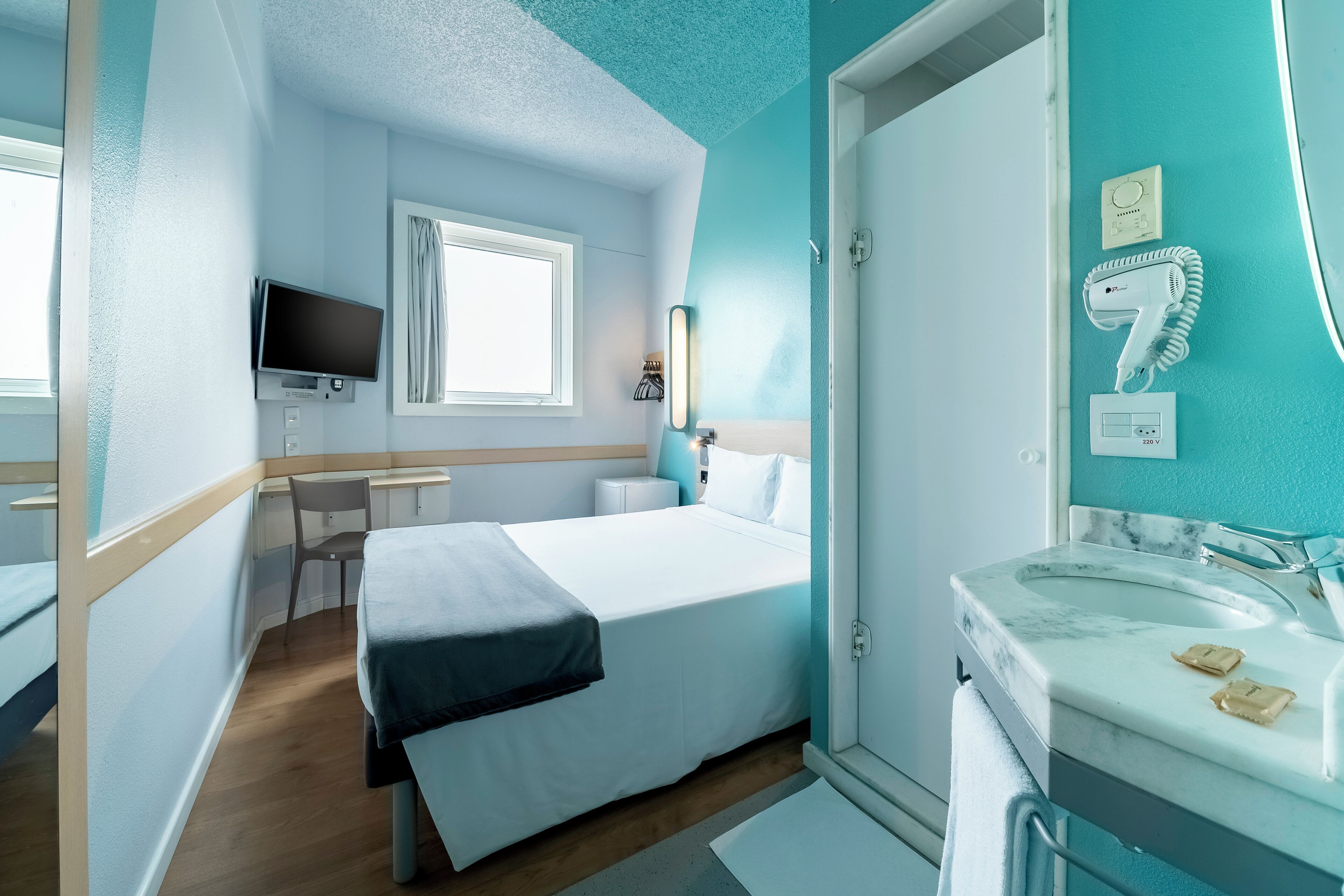 A hotel room with a bed, a desk, a TV, a window, and a bathroom with a sink and a hairdryer.