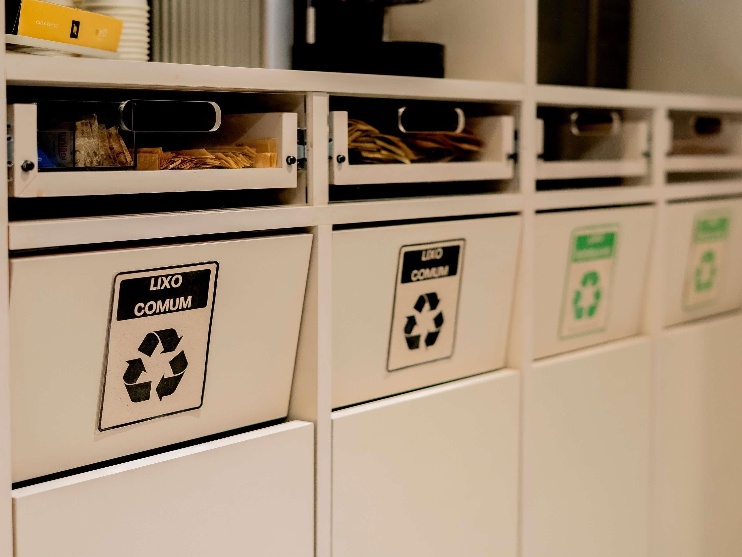 Una fila de contenedores blancos de reciclaje con etiquetas que indican 'LIXO COMUM' y símbolos de reciclaje.