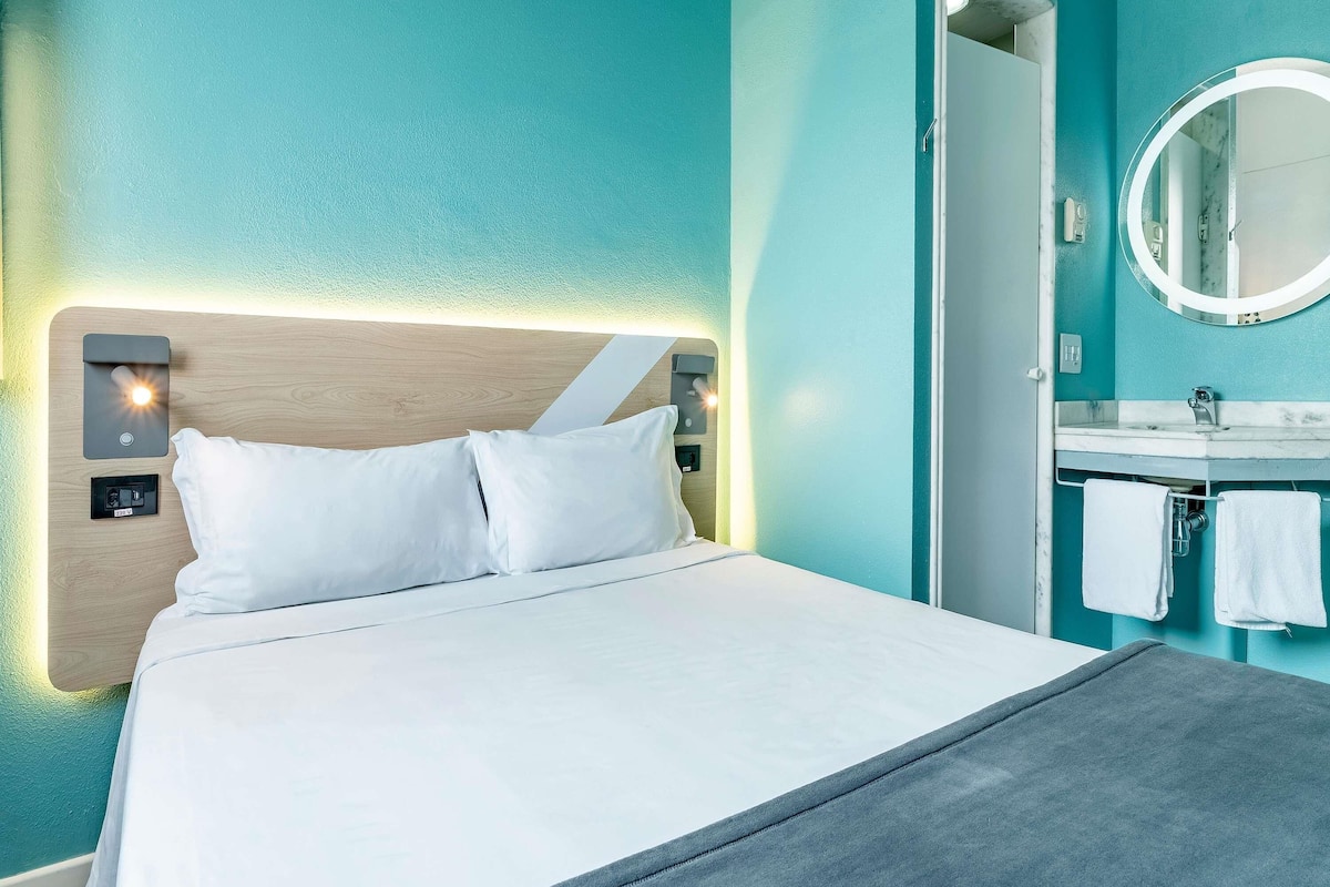A hotel room with a bed, a bedside table, a nightstand, a bathroom with a sink and mirror, and a towel rack.