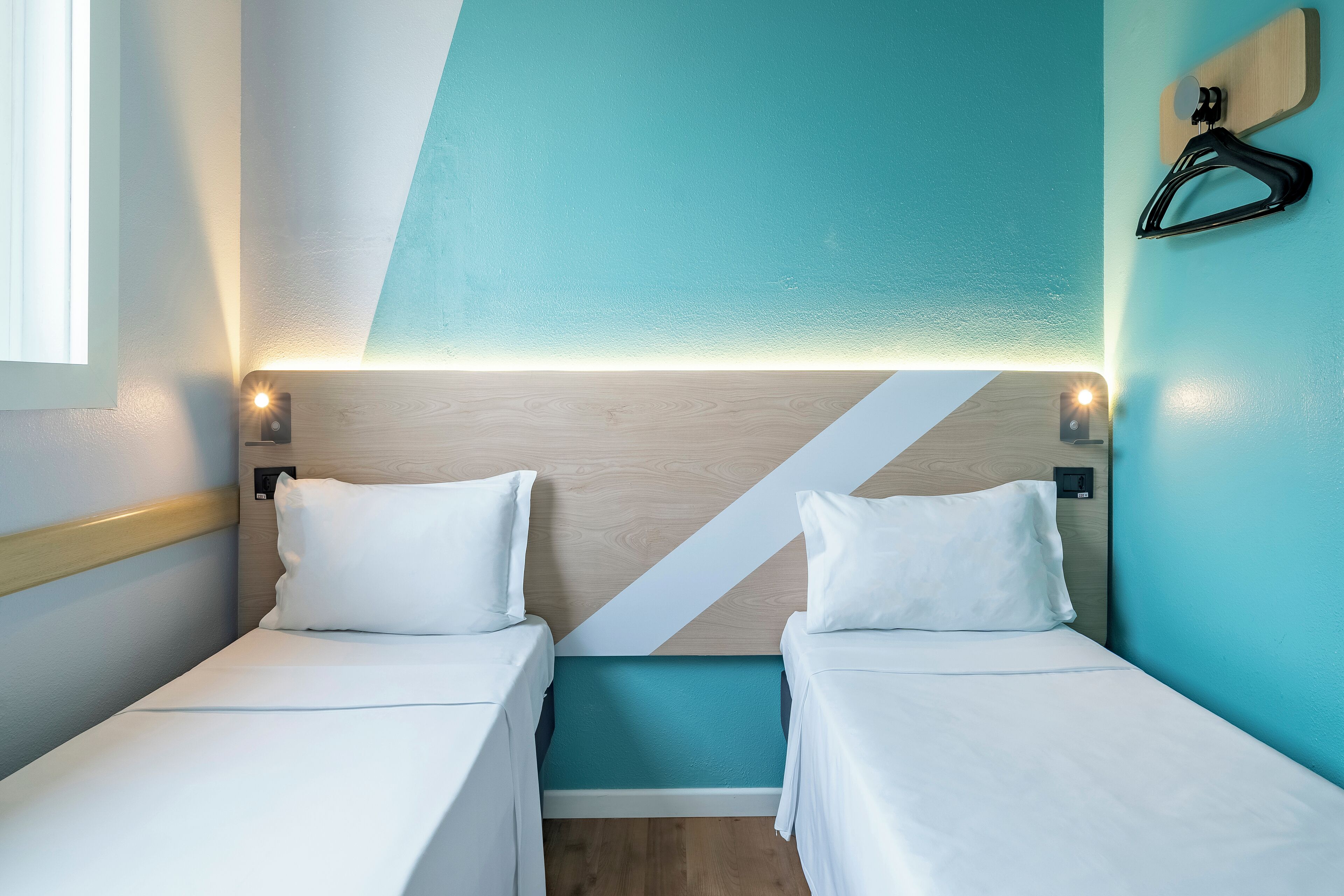A hotel room with two beds, a wooden headboard, teal accent wall, and a black hanger on the wall.