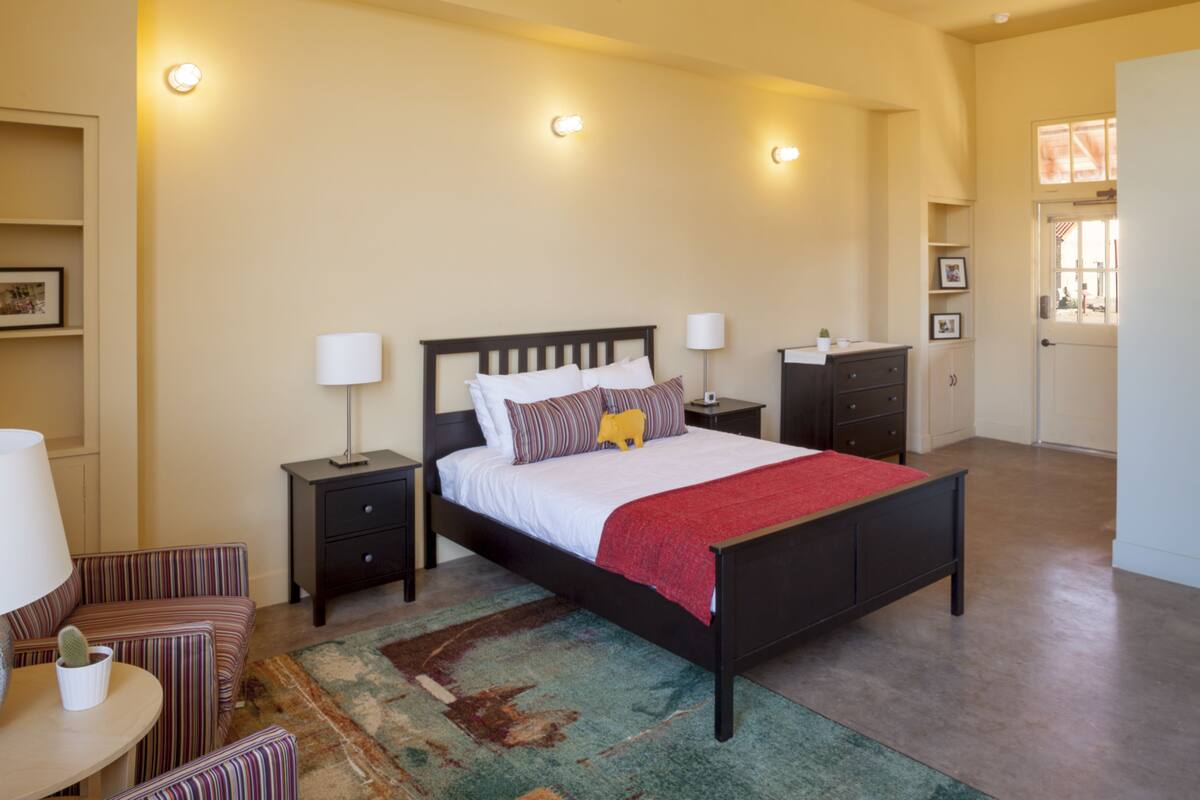 Un dormitorio con cama, mesitas de noche, lámpara, silla y alfombra.