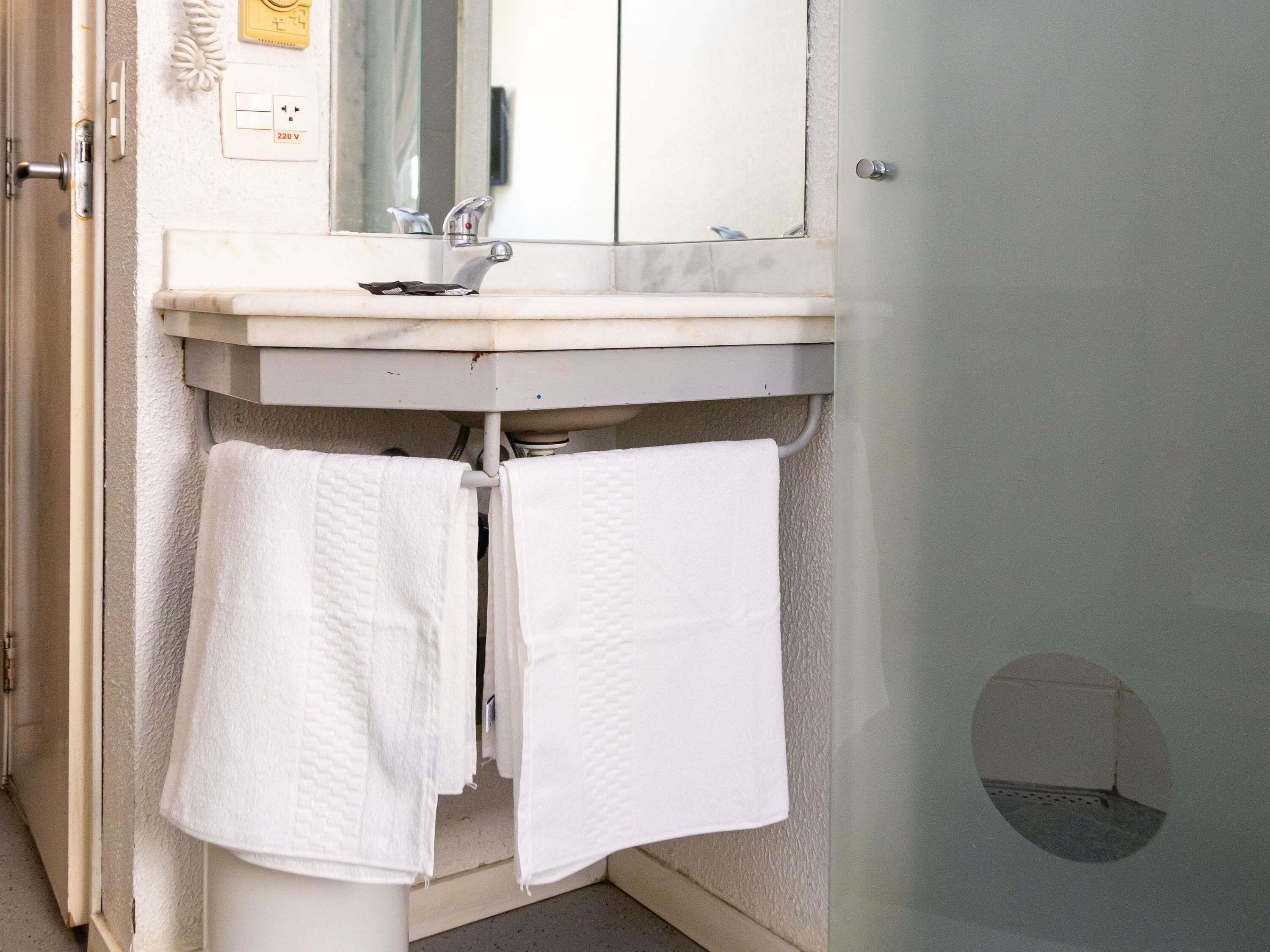 Un baño con lavamanos, espejo, toallero y un enchufe de 220 V.