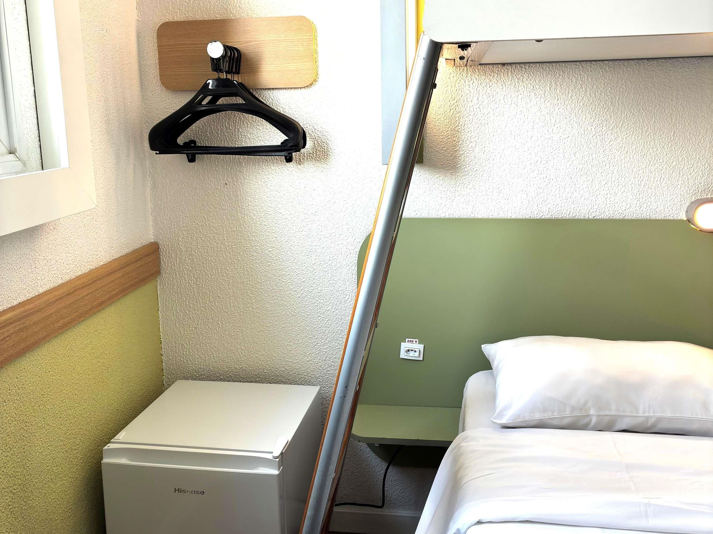 Een kleine hotelkamer met een bed, een nachtkastje, een wandhaak en een stopcontact.