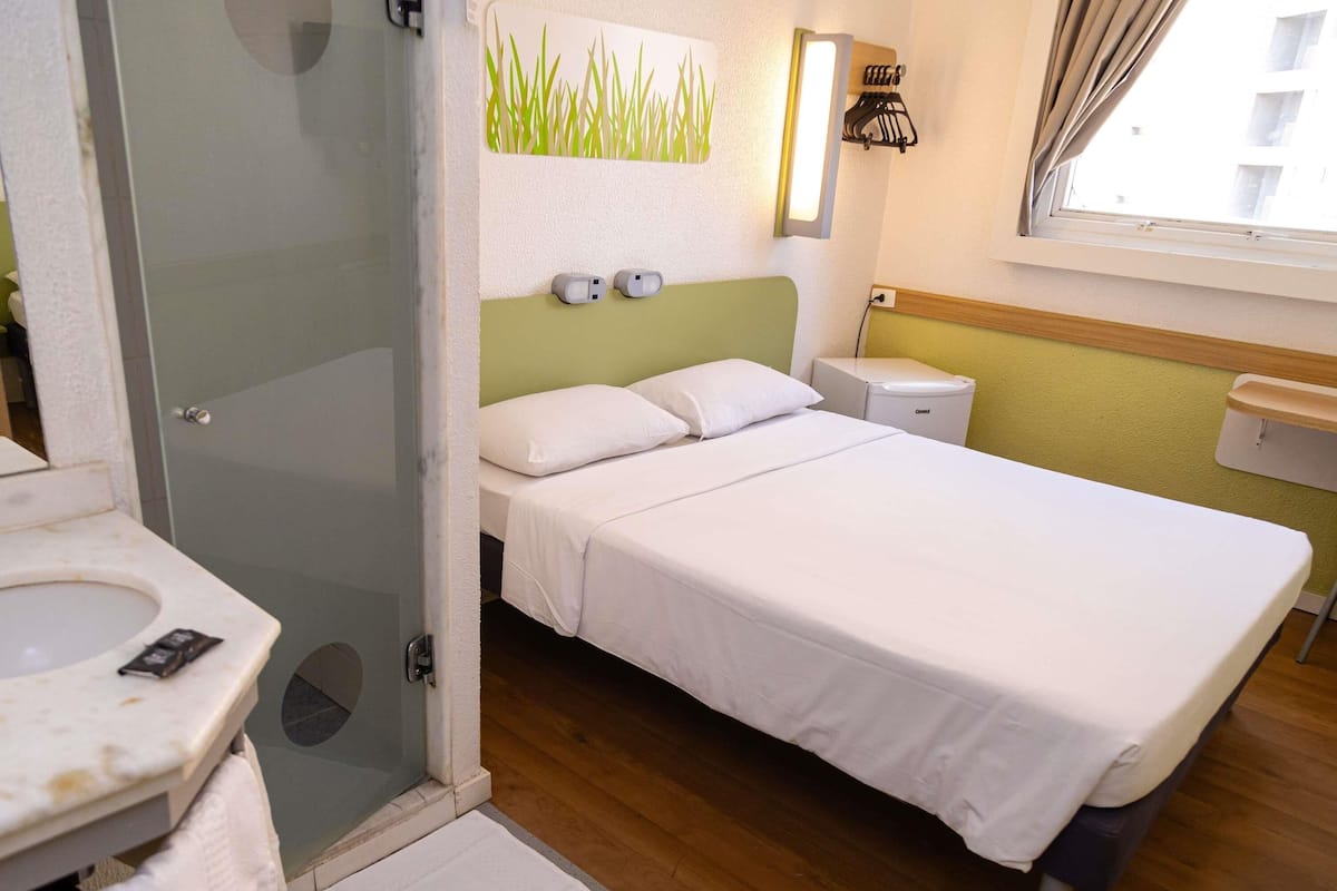 A hotel room with a bed, a bedside table, a nightstand, a mirror, a towel, a shower area, and a window with curtains.