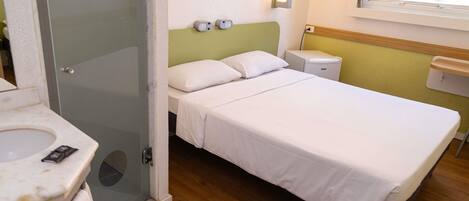 Habitación de hotel con cama, mesita de noche, espejo, toalla, zona de ducha y ventana con cortinas.