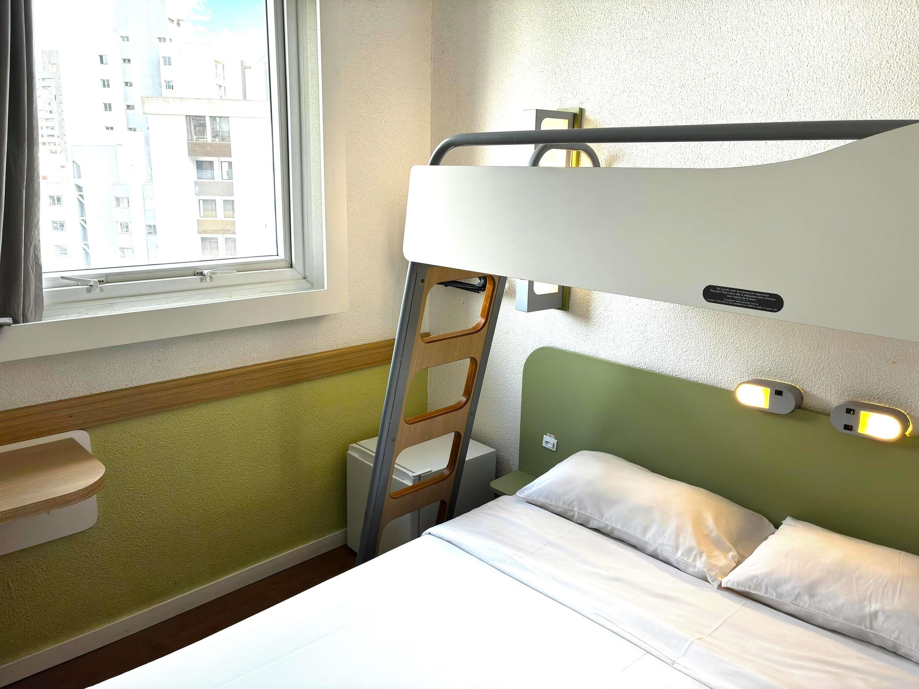 Una litera con escalera, una mesita de noche y una ventana con vista a edificios.