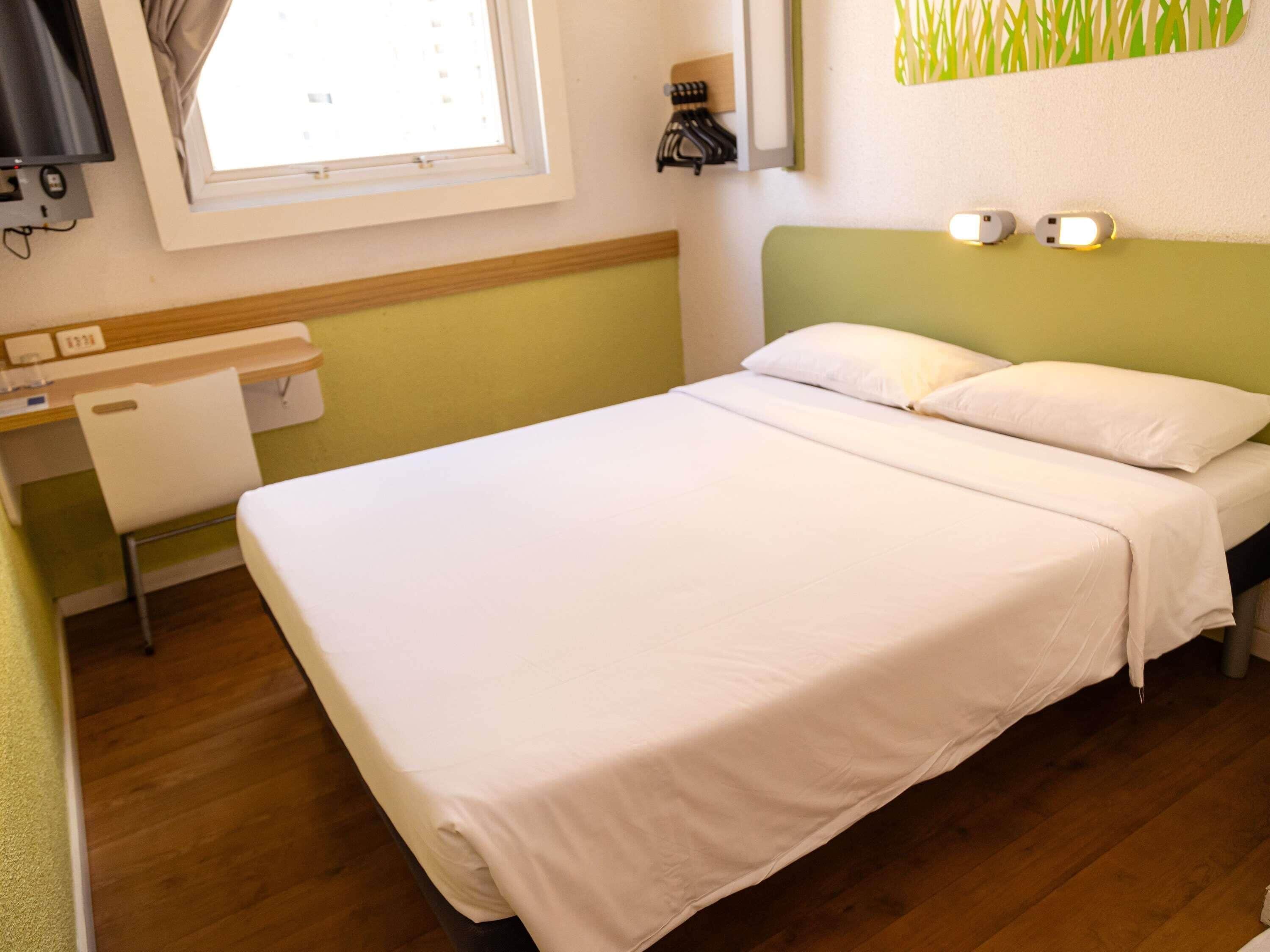 A single bed with white bedding, a desk with a chair, a window with curtains, and a wall-mounted light fixture.