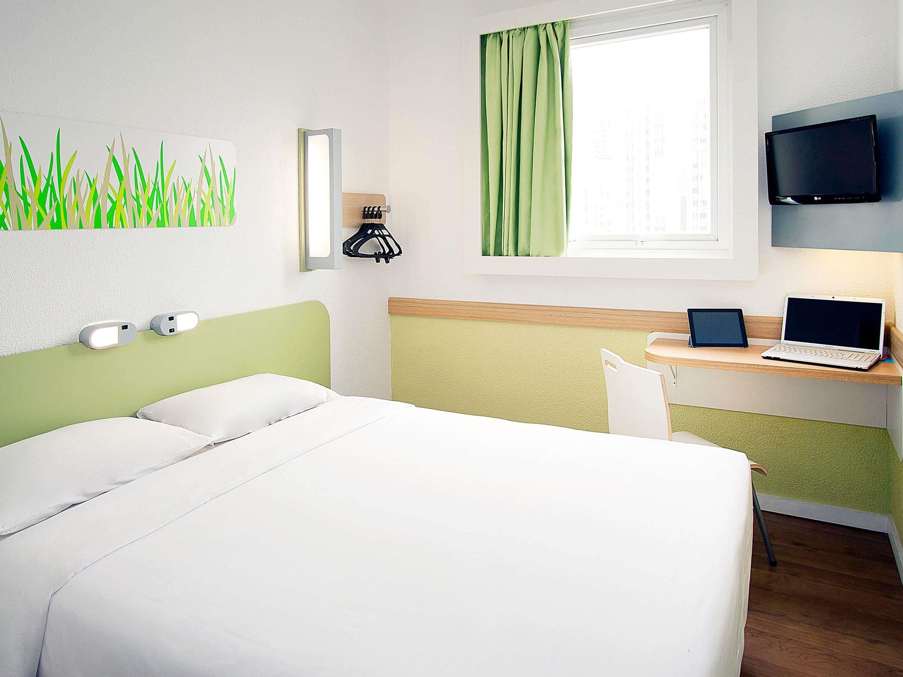 Une chambre d’hôtel équipée d’un lit, d’un bureau avec un ordinateur portable et une tablette, d’une télévision et d’une fenêtre avec des rideaux.