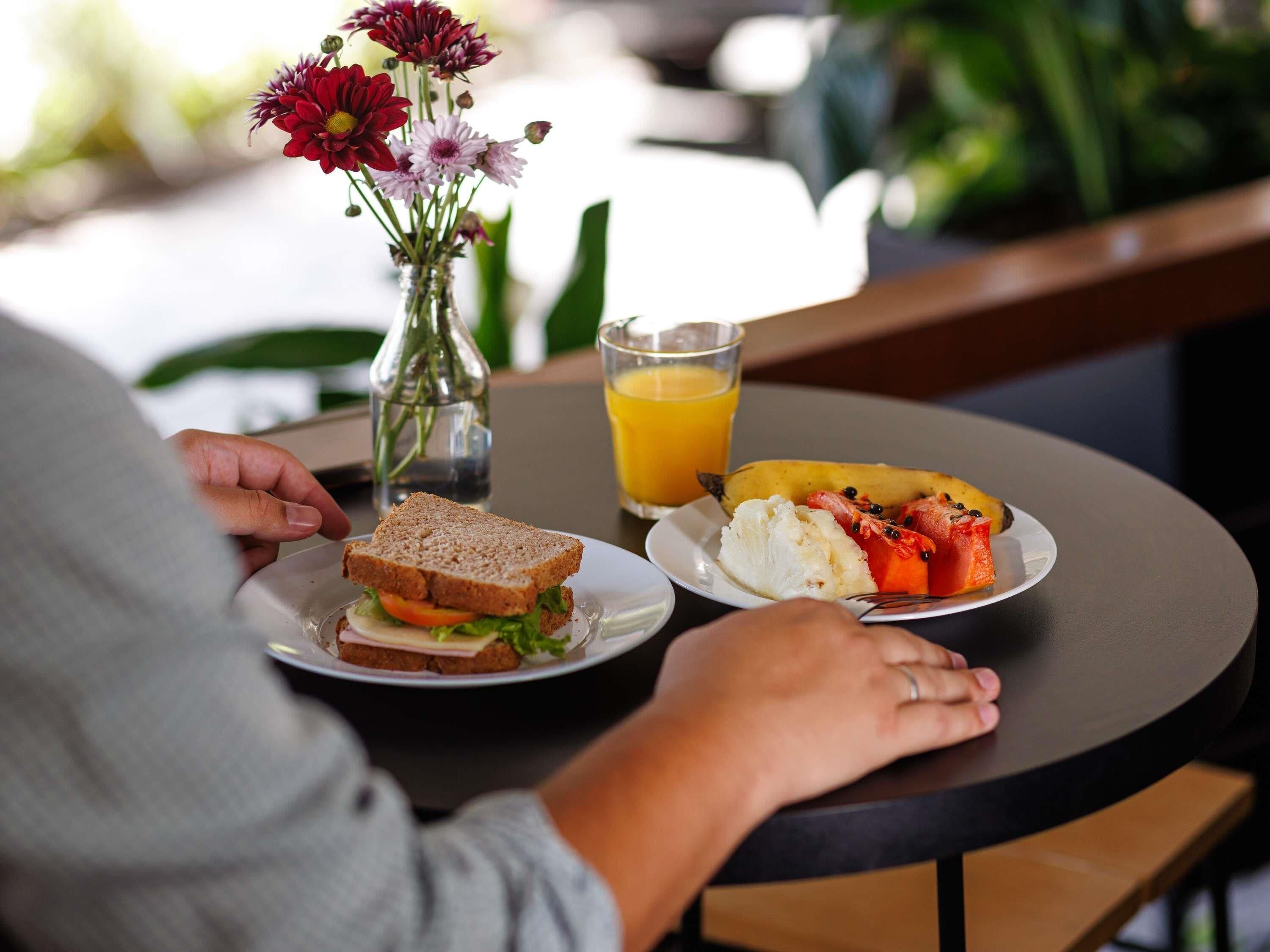 A person sitting at a round table with a sandwich, a plate of fruit, a glass of orange juice, and a vase with flowers.