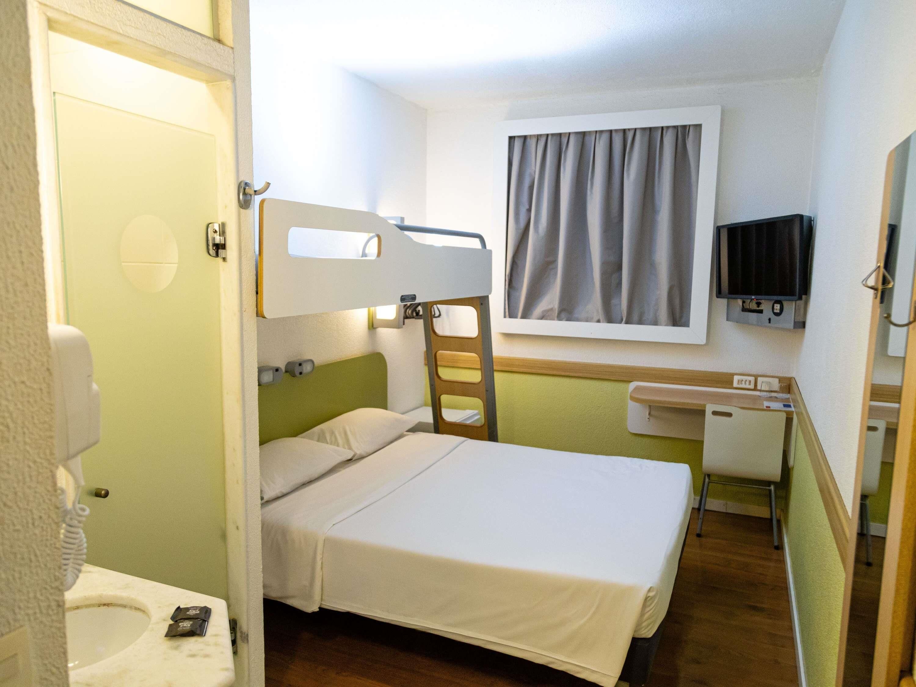 A compact hotel room with a bunk bed, a small desk, a television, and a window with curtains.