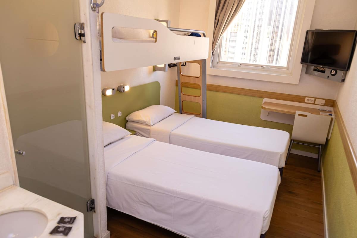 A small, compact hotel room with two single beds, a toilet, a sink, and a television mounted on the wall.
