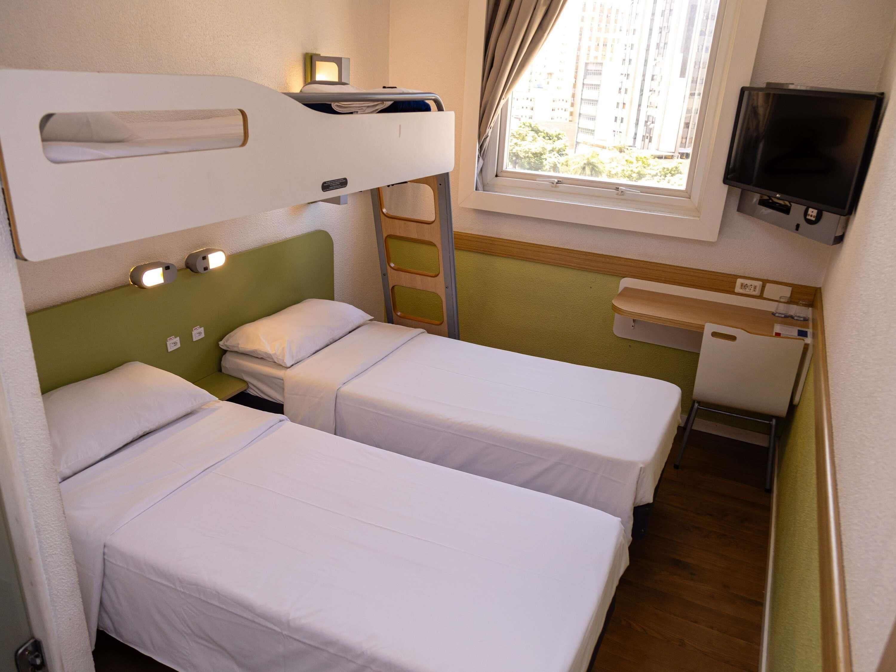 A small, compact hotel room with bunk beds, a TV, a window with curtains, and a small desk with a chair.