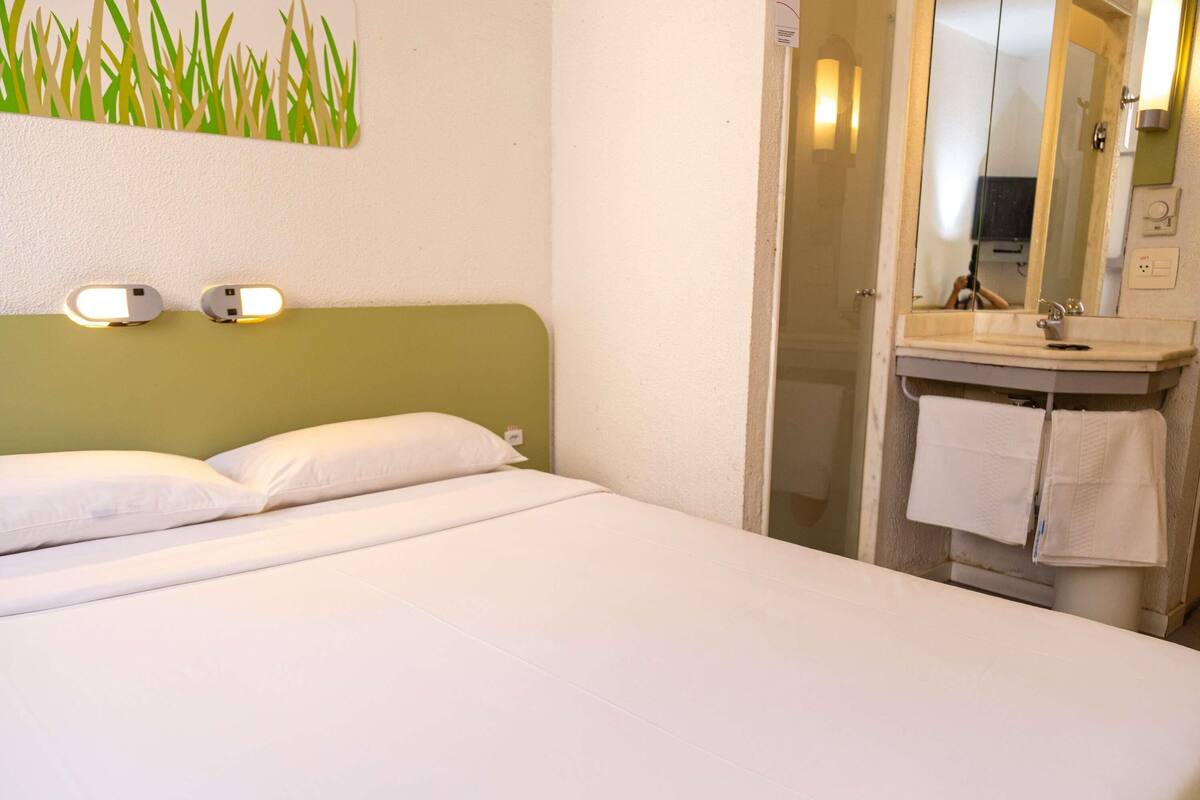 A hotel room with a bed, a green headboard, a wall-mounted light fixture, a bathroom with a mirror, and a towel rack.