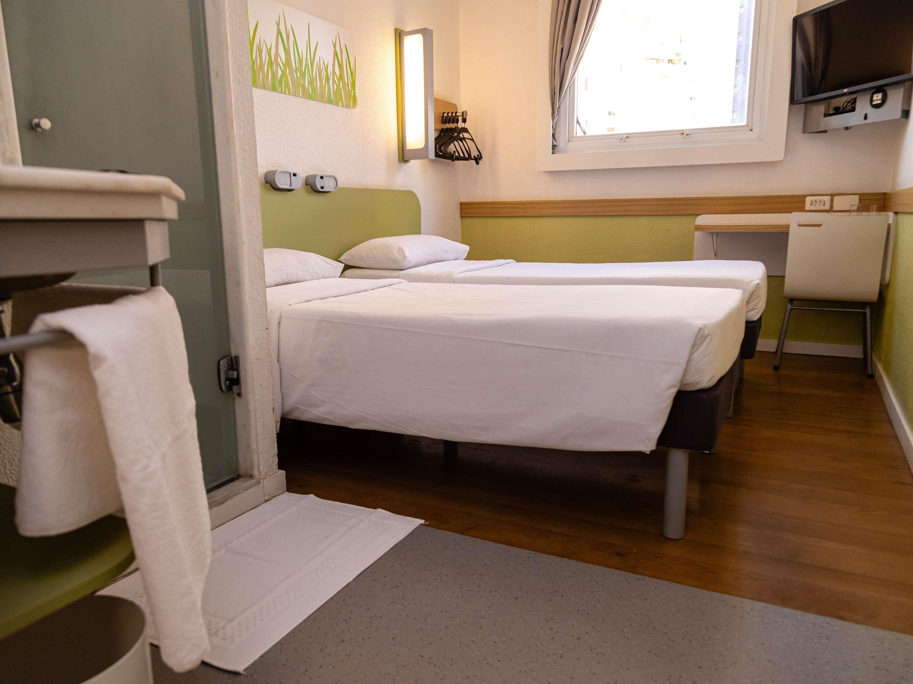 A hotel room with two beds, a desk, a television, and a window.