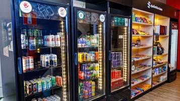 Três máquinas de venda automática de bebidas com diversas opções e uma prateleira com lanches à direita.