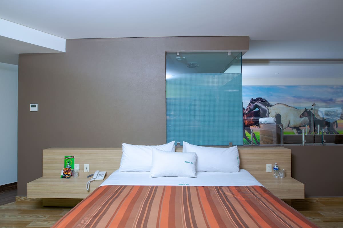 A hotel room with a bed, a desk with a phone and a brochure, a water bottle, and a painting of horses on the wall.