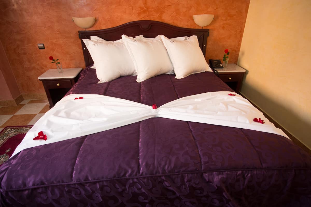 A neatly made bed with a purple bedspread and white pillows, a bedside table with a lamp, and a vase with a single red rose.