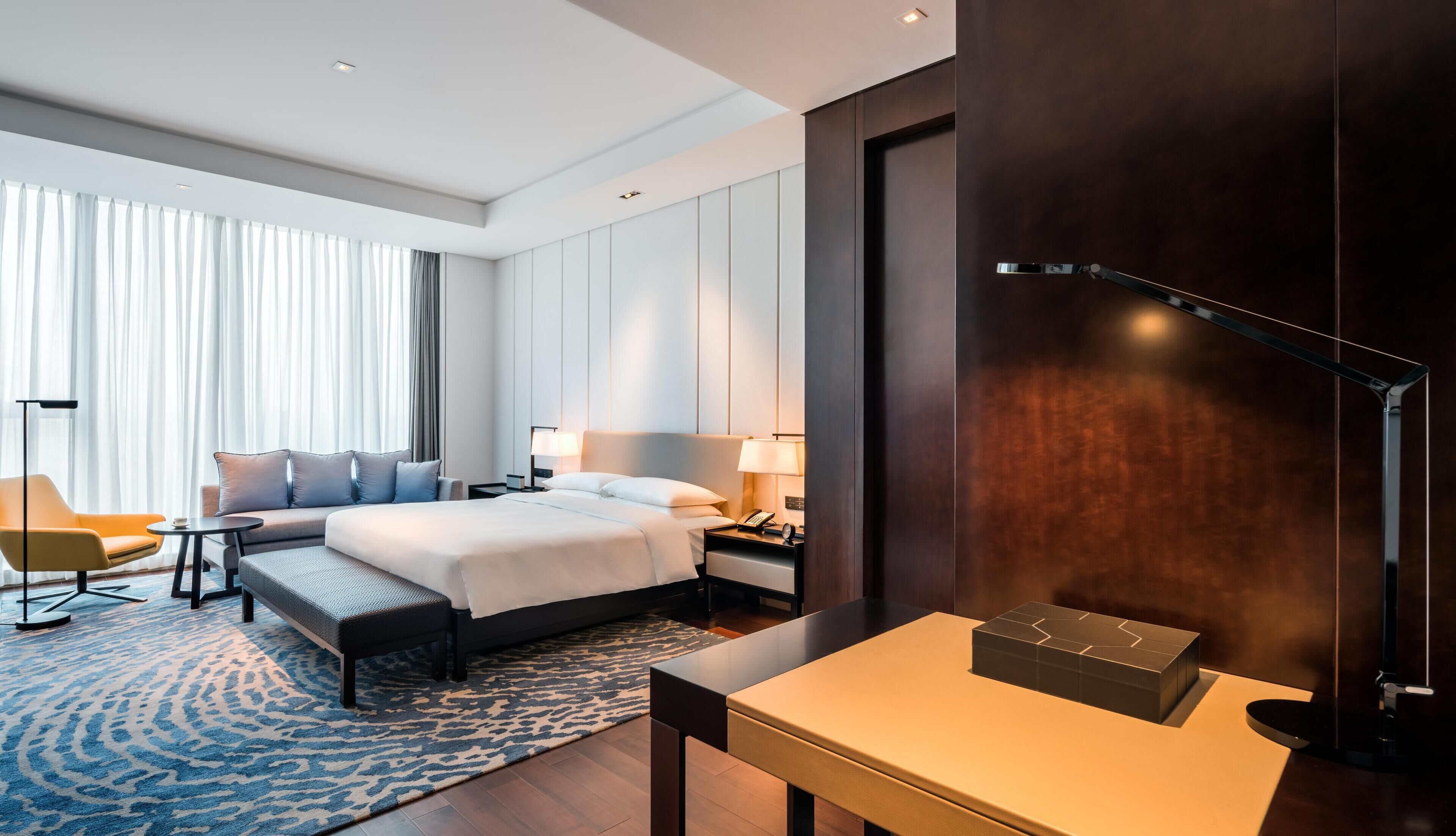 Ein Hotelzimmer mit Bett, Nachttischen, einem Schreibtisch, einem Sessel und einem großen Fenster mit Vorhängen.
