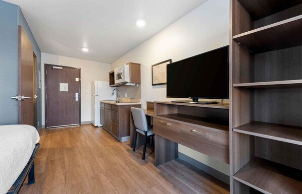 Habitación de hotel pequeña con cama, kitchenette, escritorio y estanterías integradas.