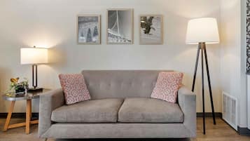 A neutral-toned sofa with patterned cushions, a small side table with a lamp and a vase of flowers, and three framed pictures on the wall.