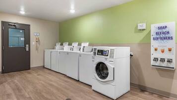 Een wasserette met meerdere wasmachines en drogers, een rookverbod bord en een poster met advertenties voor wasmiddel, bleekmiddel en wasverzachter.