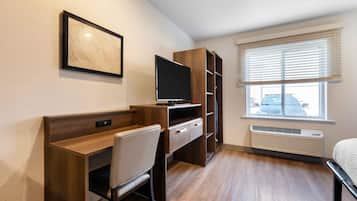 Ein kleines Hotelzimmer mit Schreibtisch aus Holz, einem Sessel, einem Fernseher und einem Fenster mit Jalousien.