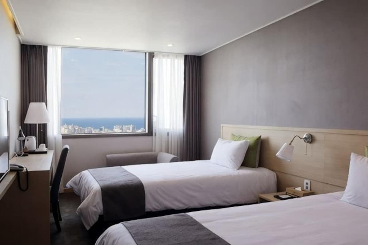 A hotel room with two beds, a desk, a chair, a lamp, and a view of the city.