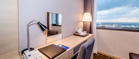 Habitación de hotel con escritorio, teléfono, espejo, lámpara, laptop y vista de la ciudad.