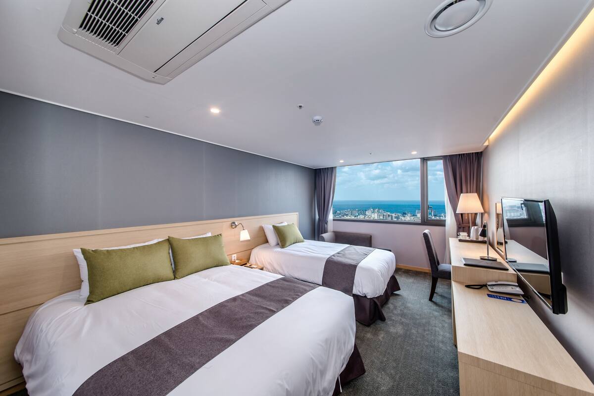 A hotel room with two beds, a desk with a computer, and a large window offering a city view.