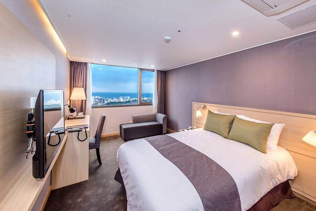 A hotel room with a bed, desk, chair, TV, and a view of the city.